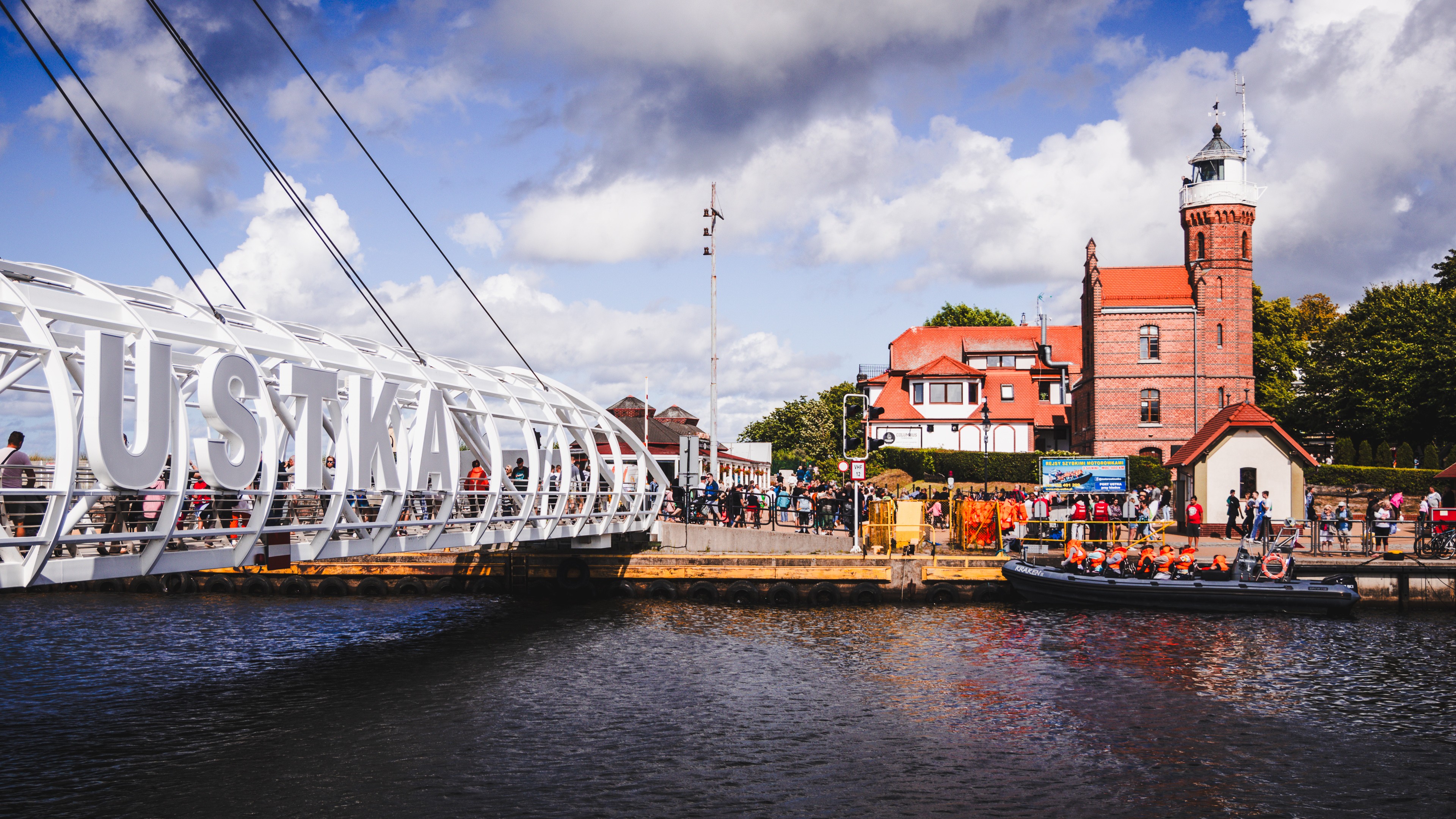Zdjęcie przedstawia nadmorską promenadę z nowoczesną kładką dla pieszych, latarnią morską i grupą ludzi